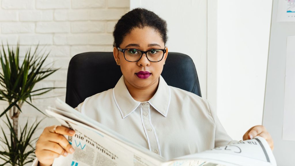 Professional woman with glasses reading a newspaper at her office desk, surrounded by modern decor. — Enhance Your Experience