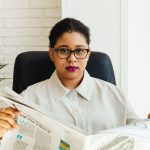 Professional woman with glasses reading a newspaper at her office desk, surrounded by modern decor. — Enhance Your Experience