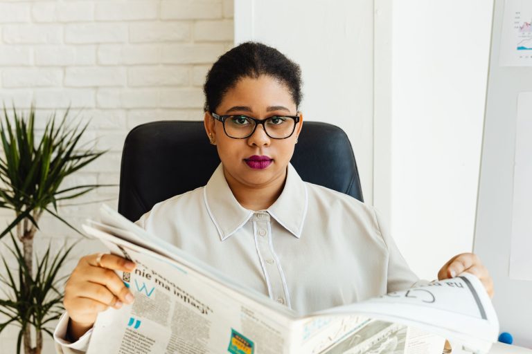 Professional woman with glasses reading a newspaper at her office desk, surrounded by modern decor. — Enhance Your Experience