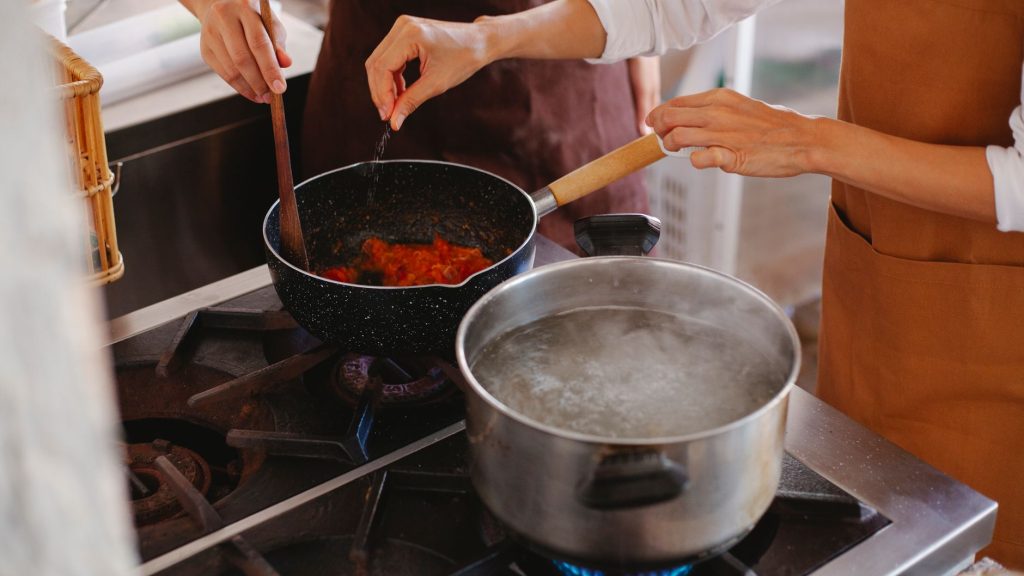 Close-up of two women cooking; stirring sauce and boiling water on stove. — Mediterranean Magic: Burst Cherry Tomato & Ro