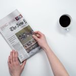 Flat lay of hands holding The New York Times newspaper with coffee on a white table. — The Legalities of Buying Weed Online